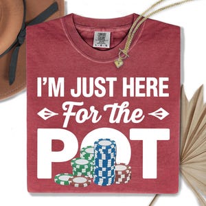 May include: A red t-shirt with the text "I'M JUST HERE For the POT" in white. The design includes stacks of poker chips in green, blue, and red. A gold necklace with a heart pendant is visible.