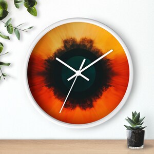 May include: A white wall clock with a circular orange, yellow, and black abstract design. The clock hands are white and black.