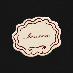 Pode incluir: Cart&atilde;o de nome creme com borda ondulada e o nome "Marianna" em escrita bord&ocirc;. O cart&atilde;o tem enfeites decorativos bord&ocirc; na parte inferior. O fundo &eacute; preto.
