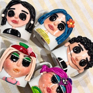 May include: Five white ceramic mugs featuring three-dimensional clay character faces. Each face has black button eyes, distinct hair colours including black, blue, blonde, curly black, and purple, and unique facial features like stitches or flowers.