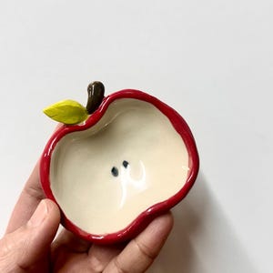 May include: A small ceramic dish shaped like a halved apple. The exterior rim is painted bright red, while the interior is a smooth off-white colour featuring two small black dots. A brown stem and a single yellow-green leaf are attached to the top.