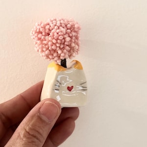 May include: A small ceramic cat-shaped planter with a cream-coloured body, yellow ears, and a red heart-shaped nose. The planter holds a miniature tree with a dense, textured canopy of tiny, light pink spherical blossoms on a dark brown trunk.