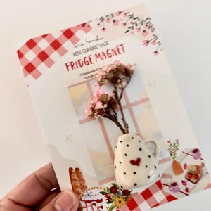 May include: A cream-coloured ceramic mini vase fridge magnet with black polka dots and a small red heart, holding dried pink flowers. The magnet is attached to a decorative card featuring a red and white gingham pattern and window illustration.