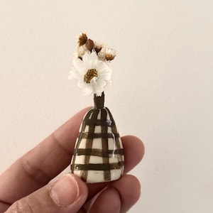 May include: A miniature ceramic vase with a glossy white finish and a dark brown grid pattern. The vase has a narrow neck and a rounded base, containing a small arrangement of dried white flowers with yellow centres and brown accents.