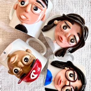 May include: Four white ceramic mugs featuring three-dimensional clay faces. Each mug displays a unique character with distinct hair colours, facial features, and clothing details, including one figure wearing a red cap with a white ampersand symbol.