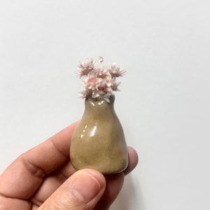 May include: A small, smooth, tan-coloured ceramic vase with a rounded, organic shape. The vase features a narrow neck and contains a cluster of tiny, delicate, light pink dried flowers that extend from the opening at the top of the vessel.