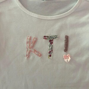 Può includere: Maglietta bianca a costine con le lettere "KT!" cucite in tessuto rosa con motivi floreali e a quadri.