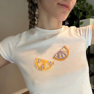 Può includere: Maglietta bianca con due fette di limone gialle applicate sul davanti. Le fette di limone sono fatte di tessuto e hanno un motivo a quadri viola e bianchi.