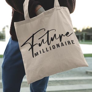 May include: A beige canvas tote bag with the words "Future Millionaire" printed in black script. The bag has two handles and is being held by a person wearing a dark sweater and blue jeans. The bag is a neutral color.