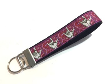 May include: A fabric wristlet keychain featuring a repeating pattern of white hand signs for I love you against a magenta and purple background. The strap is attached to a silver-toned metal clasp and a circular split ring for keys.