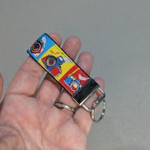 May include: A colorful camera-themed keychain. The ribbon features illustrations of vintage cameras in blue, red, and yellow squares. The keychain has a black fabric end and a silver metal ring.