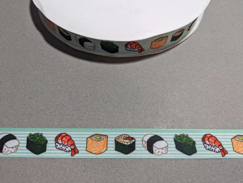 May include: A spool of light green grosgrain ribbon featuring a repeating pattern of various sushi rolls and shrimp nigiri. The ribbon has a textured, ribbed surface with a consistent horizontal orientation of the colorful food illustrations.