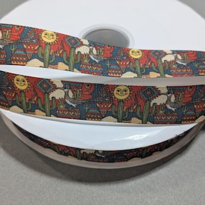 May include: Decorative ribbon featuring a Southwestern design with cacti, smiling suns, and birds. The ribbon is primarily red, blue, and beige, and is wrapped around a white spool. The ribbon is suitable for crafts and gift wrapping.