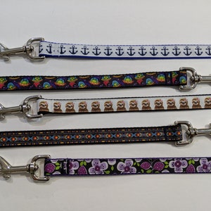 May include: Five different dog leashes with various colorful patterns. The leashes are all black with different colored patterns. The patterns include anchors, sloths, geometric designs, and flowers.
