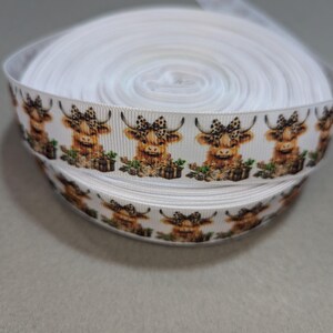 May include: White ribbon with a repeating pattern of brown and white Highland cows wearing bows. The cows are in a sitting position and have a brown and white spotted pattern.