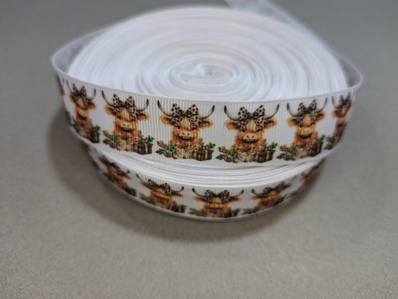May include: White ribbon with a repeating pattern of brown and white Highland cows wearing bows. The cows are in a sitting position and have a brown and white spotted pattern.