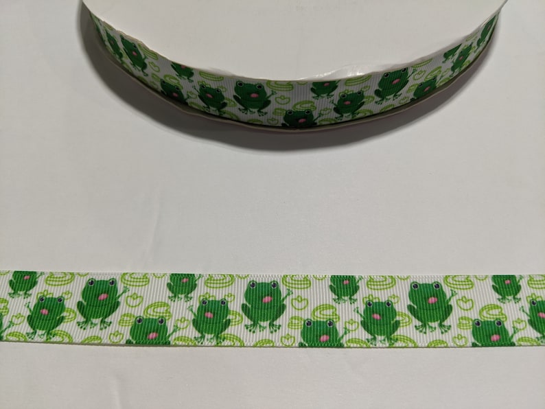 May include: White ribbon with a repeating pattern of green frogs with pink tongues on a white background.