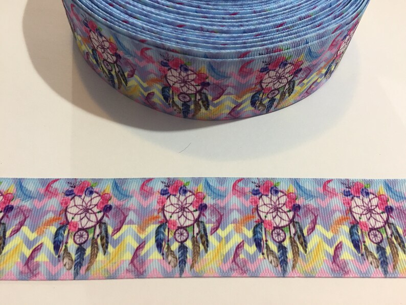Pu&ograve; includere: Un rotolo di nastro blu con un motivo colorato di acchiappasogni. Il nastro presenta uno sfondo a chevron con accenti rosa, gialli e blu. Gli acchiappasogni sono circondati da piume e fiori.
