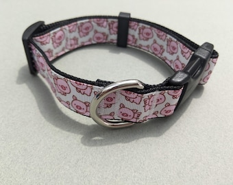 May include: A white dog collar with a pink pig pattern and black accents. The collar has a silver metal D-ring and a black plastic buckle.