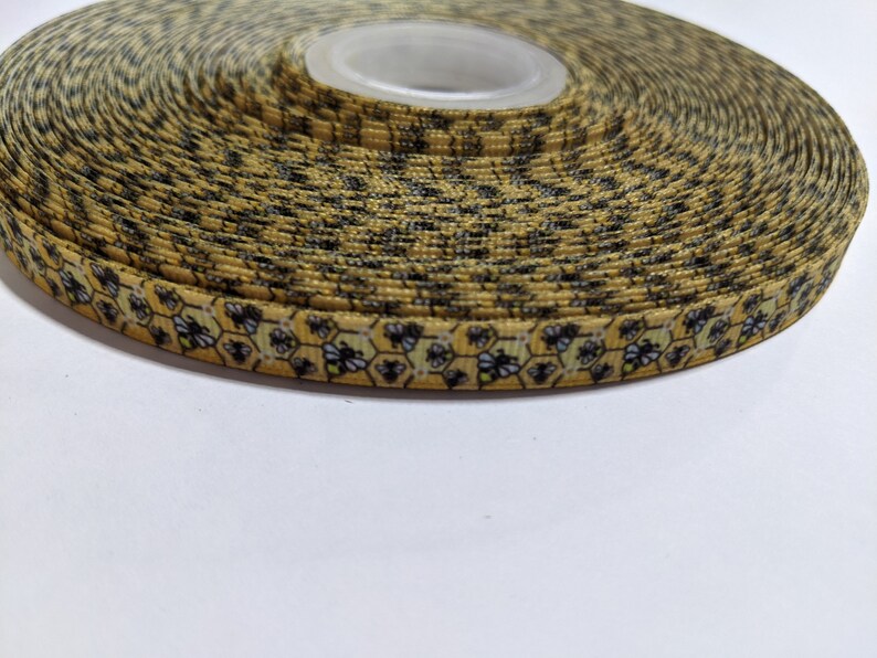 May include: A roll of patterned ribbon featuring a bee and honeycomb design on a yellow background. The ribbon is tightly wound, displaying the repeating pattern. Suitable for crafts, gift wrapping, or decorative applications. The ribbon is likely measured in meters.