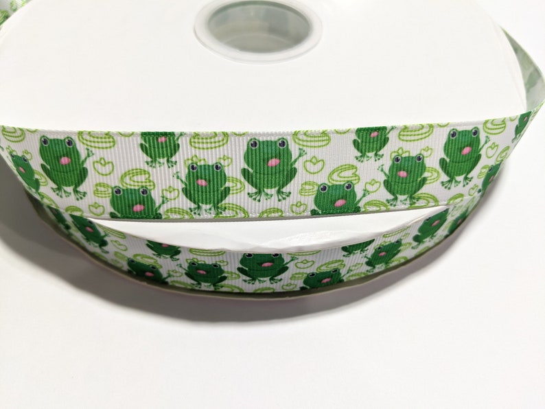 May include: A roll of white ribbon with a green frog pattern. The ribbon is on a white spool.