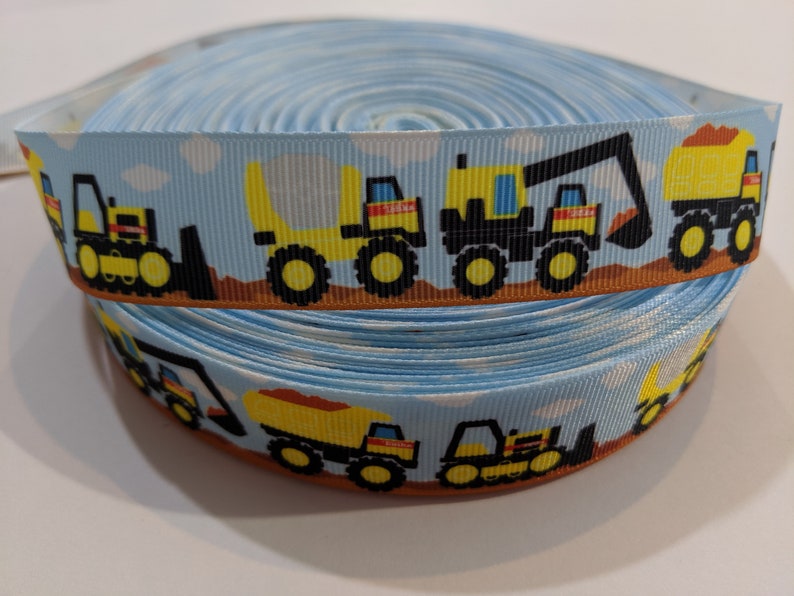 May include: A roll of blue ribbon with a repeating pattern of yellow construction vehicles on a brown background. The vehicles include a dump truck, an excavator, and a bulldozer.