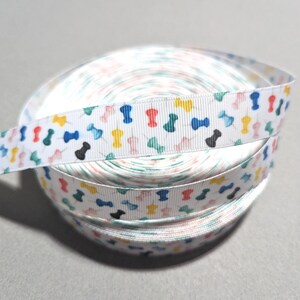 May include: A white grosgrain ribbon roll featuring a repeating pattern of small, colorful push pins in shades of red, blue, yellow, green, black, and pink. The ribbon has a textured, ribbed surface and is wound into a tight, circular spool.