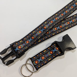 May include: A black lanyard featuring a repeating geometric pattern in shades of blue, orange, and yellow. The strap includes a black plastic side-release buckle and a detachable key fob section with a matching buckle and two silver metal rings.
