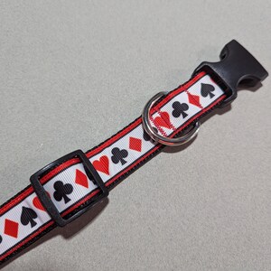 May include: A white, red, and black dog collar with a repeating pattern of playing card suits. The collar has a black plastic buckle and a silver metal D-ring.