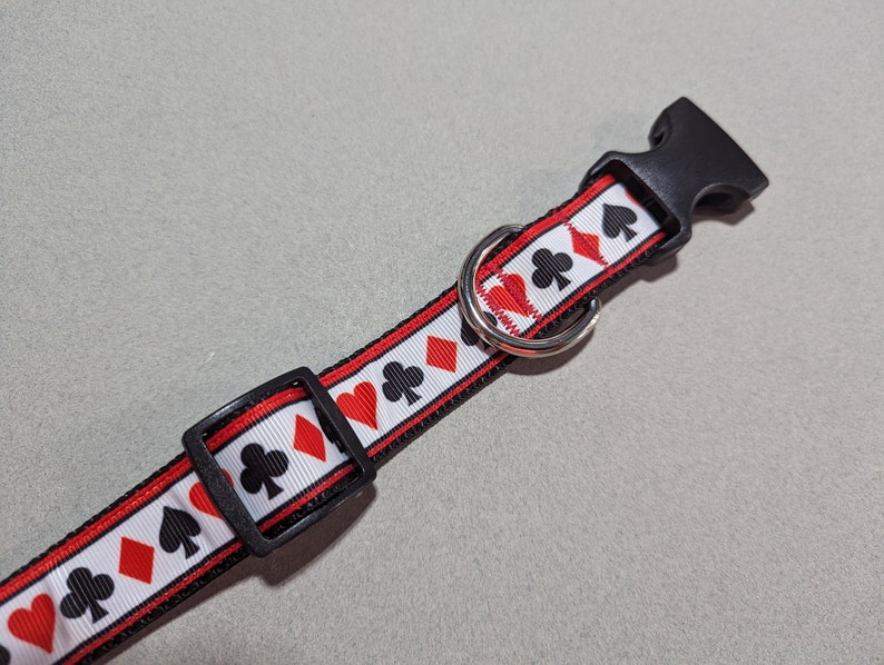 May include: A white, red, and black dog collar with a repeating pattern of playing card suits. The collar has a black plastic buckle and a silver metal D-ring.