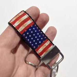 May include: A black and red key chain with a distressed American flag design. The key chain has a silver metal ring and a silver metal clasp.