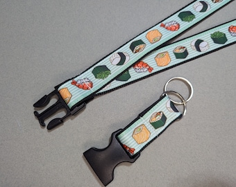 May include: A black and white lanyard with a green and white pattern of sushi rolls. The lanyard has a black plastic clip and a silver key ring.
