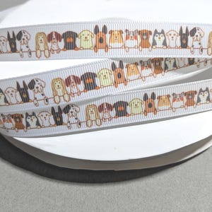 May include: A white grosgrain ribbon wound on a spool, featuring a repeating pattern of various dog breeds in brown, black, tan, and white colors. The dogs are illustrated in a line, appearing as if they are peeking over a horizontal fence line.