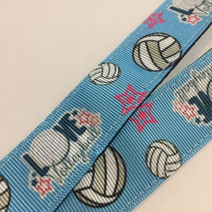 May include: Light blue lanyard with a repeating pattern of volleyballs, red stars, and the word "Volleyball" in white. The lanyard is made of a ribbed fabric.