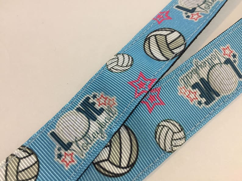 May include: Light blue lanyard with a repeating pattern of volleyballs, red stars, and the word "Volleyball" in white. The lanyard is made of a ribbed fabric.