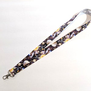 May include: A black and white lanyard with a space-themed print featuring planets, stars, and spaceships. The lanyard has a silver metal clip.