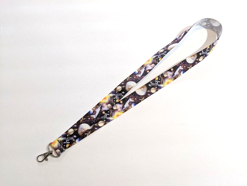 May include: A black and white lanyard with a space-themed print featuring planets, stars, and spaceships. The lanyard has a silver metal clip.