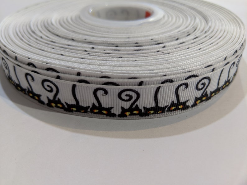 May include: White ribbon with a black cat design. The cats have curled tails and yellow eyes.