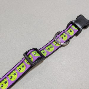 May include: A purple and black dog collar featuring a repeating pattern of green alien faces. The collar has a black plastic buckle and a metal D-ring for leash attachment. The collar is designed for pets.