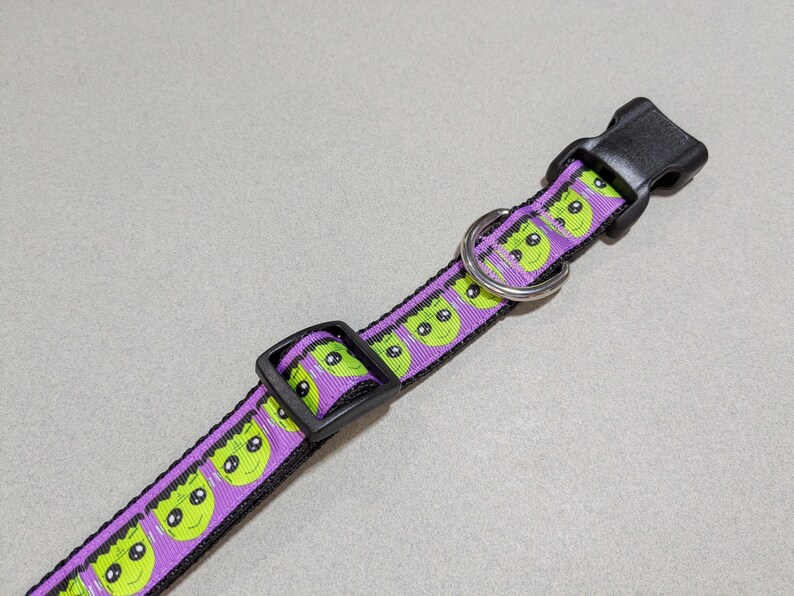 May include: A purple and black dog collar featuring a repeating pattern of green alien faces. The collar has a black plastic buckle and a metal D-ring for leash attachment. The collar is designed for pets.
