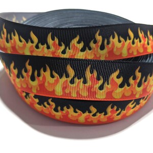 May include: A roll of black grosgrain ribbon featuring a repeating flame pattern in vibrant shades of yellow, orange, and red. The fabric ribbon has a textured, ribbed surface and is coiled into a circular shape, displaying the fiery graphic design.