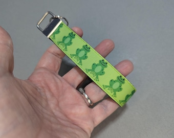 May include: A lime green wristlet keychain featuring a repeating pattern of smiling green frogs. The keychain has a silver-colored metal clasp and a black strap. The wristlet is held in a hand.