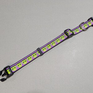 May include: A purple pet collar with a repeating pattern of green alien faces. The collar has black plastic buckles and a metal D-ring. The collar is approximately 1 inch wide.