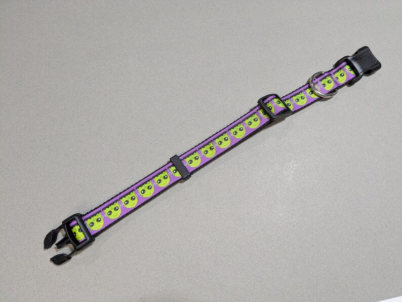 May include: A purple pet collar with a repeating pattern of green alien faces. The collar has black plastic buckles and a metal D-ring. The collar is approximately 1 inch wide.