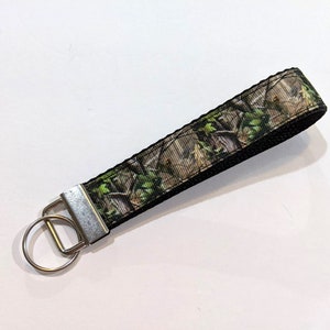 May include: A camouflage patterned key chain with a silver key ring. The key chain is made of black and green fabric with a camouflage pattern.