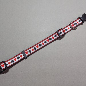 May include: A black and red dog collar with a white background and a repeating pattern of playing card suits. The collar has a black plastic buckle and a silver metal D-ring.