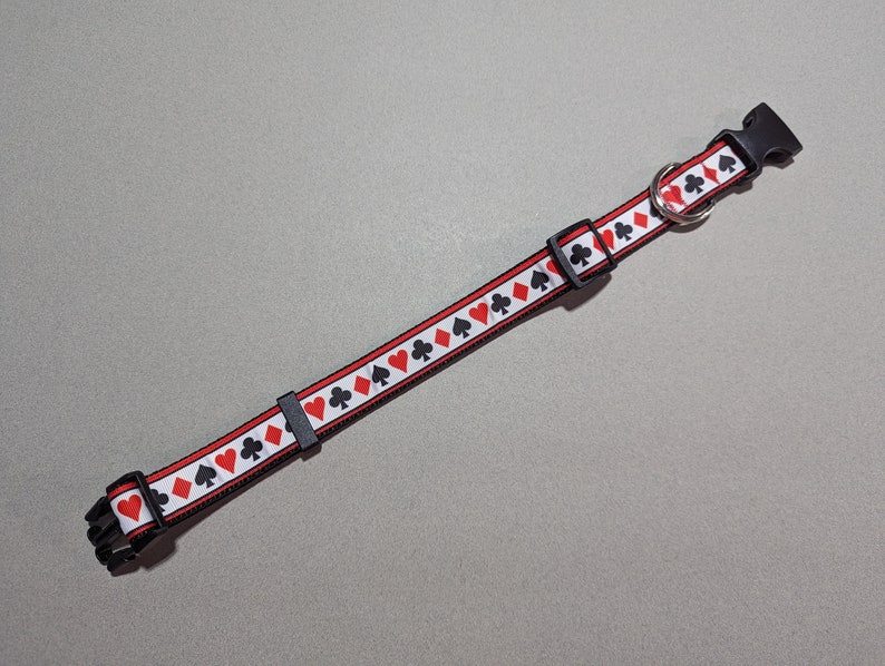 May include: A black and red dog collar with a white background and a repeating pattern of playing card suits. The collar has a black plastic buckle and a silver metal D-ring.