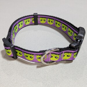 May include: A black dog collar with a purple and green monster face pattern. The collar has a black plastic buckle and a silver metal D-ring for leash attachment. The collar is designed for pets.