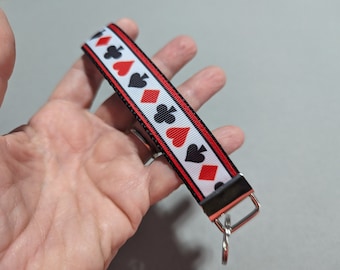 Op de afbeelding: Een rode, witte en zwarte lint sleutelhanger met een herhalend patroon van speelkaartsymbolen. De sleutelhanger heeft een zilveren metalen sleutelring.