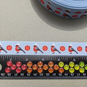 May include: A blue ribbon with a repeating pattern of red robins wearing white hats and red snowflakes. The ribbon is shown next to a ruler with inch markings.